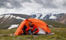 A man in an orange tent camps in an open field as stormy clouds loom over snowy mountains in the distance.