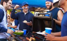 A man uses tongs to take food off of an outdoor barbecue grill. Another man stands nearby and others stand around a table.