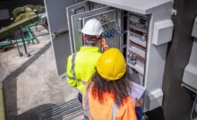 Two people wearing hard hats and safety jackets stand in front of an electrical enclosure with a clipboard.