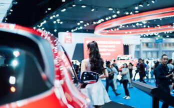 A woman in a white dress walks past a red vehicle at a car show inside a large facility with other people in the background.