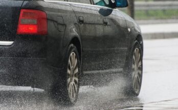 A close-up view shows a black vehicle driving down the road in a rainstorm. The tires are kicking up water.