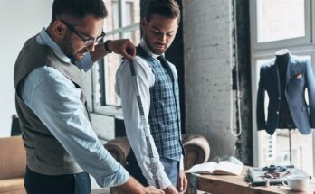 A tailor using measuring tape on the right arm of a man standing in front of him wearing a white shirt and a blue vest.