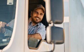 A smiling truck driver holds onto the steering wheel with one hand. He wears a baseball cap and a blue shirt.