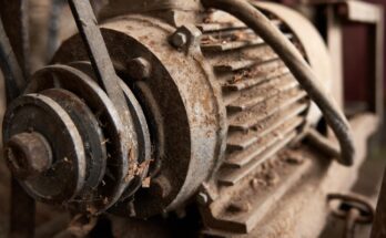 A broken electric motor with dust and other debris covering it. There is rust on the metal components of the motor.