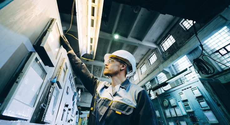 A close-up of a male power plant worker, wearing safety gear, assisting and maintaining a machine in a factory setting.