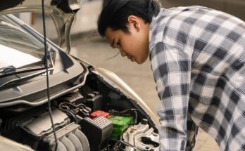 An Asian man is outside, closely examining the engine of a white car with the hood open. He's working on the car.