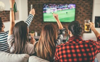 A group of four friends is sitting on the couch, cheering and drinking beverages, while watching soccer.