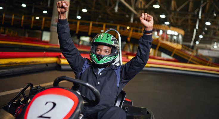 A young Black man wearing a helmet raises his fists in excitement as he sits in a go-kart on a racetrack.