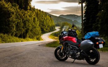A red and black motorcycle is parked at the top of a hill with a winding road below. Trees and homes align the road.