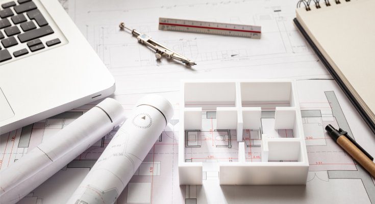 A 3D floor plan model sits on top of blueprints laid out on a table near a laptop, notepad, and drafting tools.