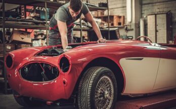A car mechanic hovering over the top of the front engine with his hand down in the compartment. The car is red.
