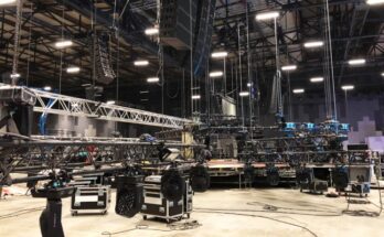 A large concert venue contains multiple lights and speakers rigged to the ceiling and road cases scattered across the floor.