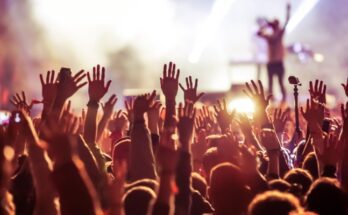 A view from the crowd at a live music concert shows people's hands raised as they vibe to music being played on stage.