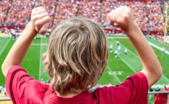 A boy wearing a red shirt raises his hands above his head to cheer on his team at a professional football game.