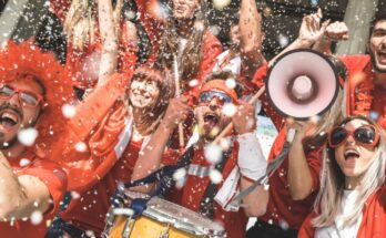 A group of friends stand near each other as they cheer on their team. Confetti flies in the air around them.