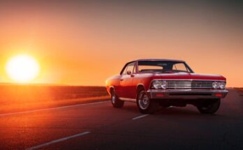 A red classic car parks sideways in the middle of an empty road. The sun is setting on the horizon in the background.