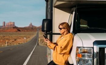 A middle-aged woman wearing sunglasses leans against a white RV on the side of the road while holding a phone.