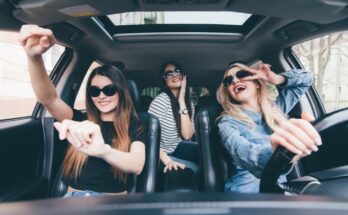 Three smiling women are in a black interior vehicle, having a fun time. One is driving, and the others are sitting.