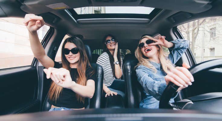 Three smiling women are in a black interior vehicle, having a fun time. One is driving, and the others are sitting.