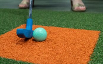A blue putter about to hit a blue golf ball on a patch of green synthetic turf with an orange square in the middle.