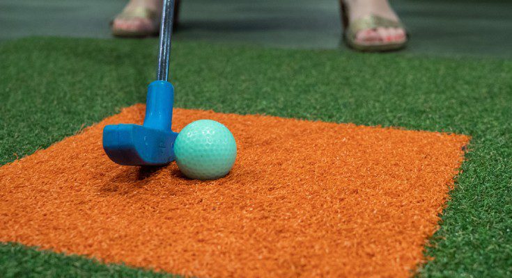 A blue putter about to hit a blue golf ball on a patch of green synthetic turf with an orange square in the middle.