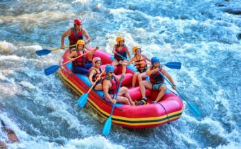 Seven people in life jackets paddle a large red raft down a rushing river during a summer whitewater rafting trip.