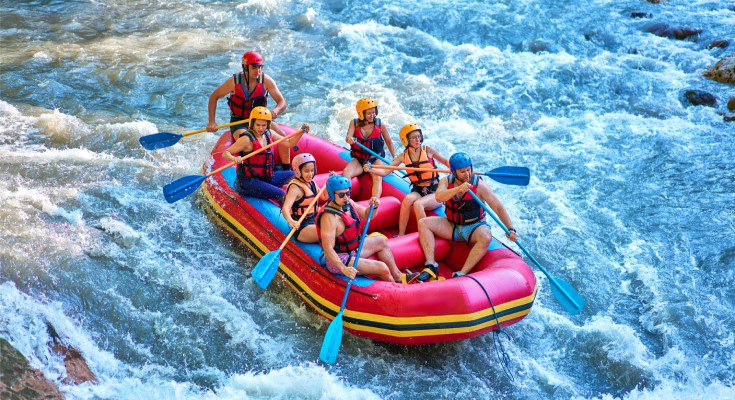 Seven people in life jackets paddle a large red raft down a rushing river during a summer whitewater rafting trip.