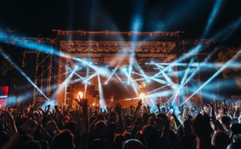 A silhouetted crowd raising their hands in front of a stage at a music festival, spotlights crisscrossing outward.