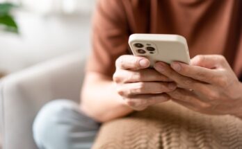 A close-up view shows a person sitting on a couch holding an iPhone in their hands. They are scrolling.