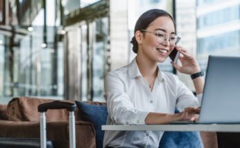 A woman sits in a lounge area with her luggage. She talks on the phone and has a laptop open on the table in front of her.