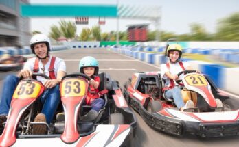 A man and a child ride together in a go-kart, driving side-by-side with a woman on an outdoor racetrack.