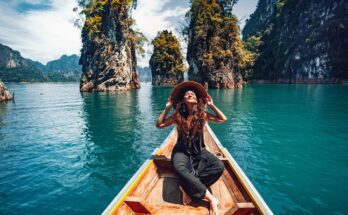 A woman dressed in a black outfit and a large hat enjoys the sun on a boat near rocky formations on the water.