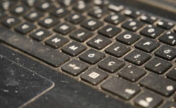 A close-up of a laptop keyboard. The keyboard has visible dust particles covering every key and the keypad.
