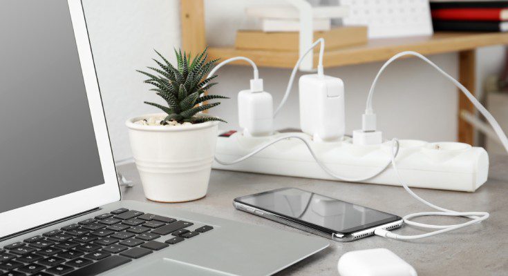 A laptop, a smartphone, a potted plant, and an AirPod case sit on a table next to a white surge protector with things plugged in.