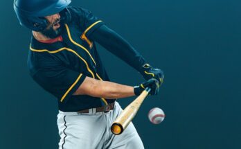An action shot of a baseball batter in a black and white jersey top with grey pants swinging a wooden bat at a ball.