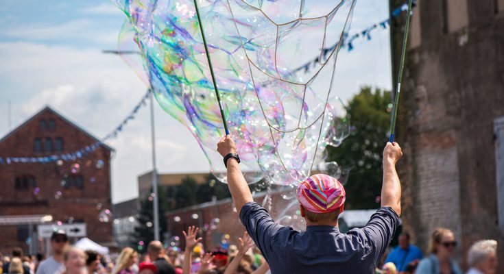 Someone is creating giant soap bubbles with rods, entertaining a crowd of children and families at an outdoor festival.