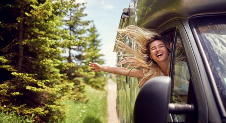 A woman leaning out of a van window on a sunny road trip, smiling and yelling with her hair blowing in the wind.