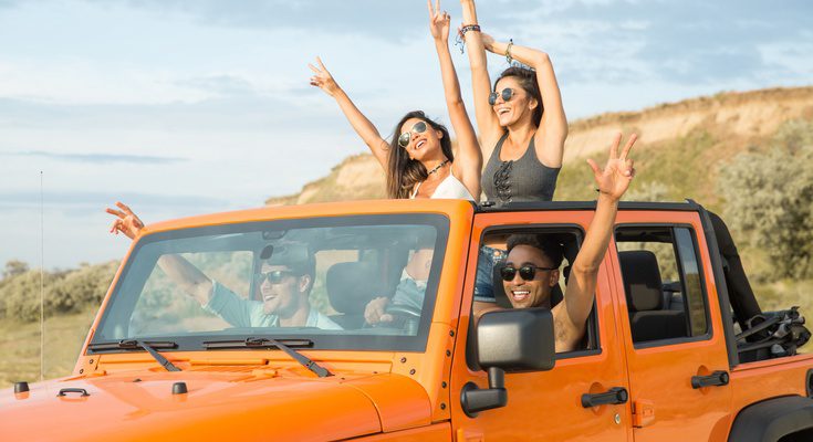 Friends celebrate in an orange Jeep during a desert off-road trip, with two women standing through the open sunroof.