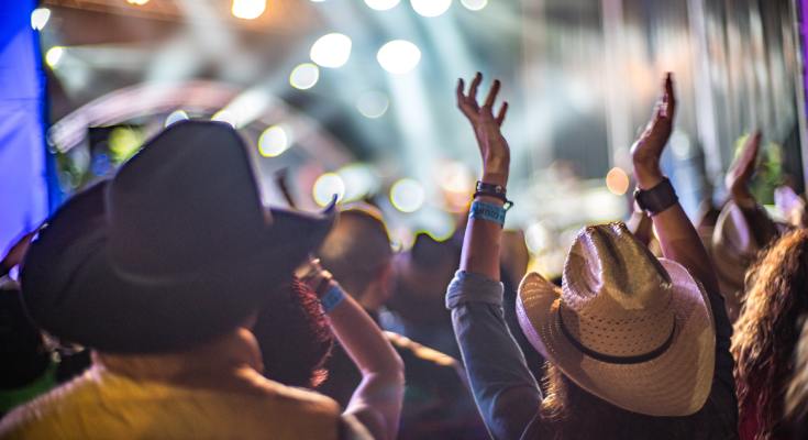 A crowd of people gathered outdoors at a country music festival, with attendees wearing hats and watching a live performance.