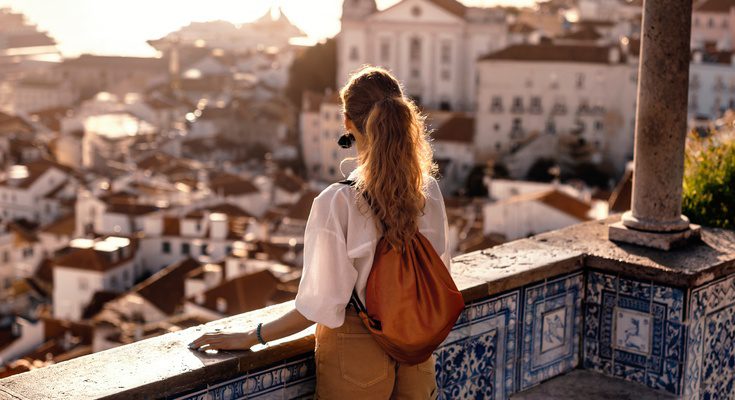 A female traveler stands at the edge of an overlook with a backpack and a ponytail. She's watching the sunset.