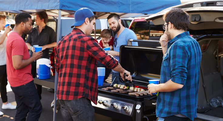 Friends stand under a canopy while holding drinks and chatting together at a tailgate beside an open SUV.