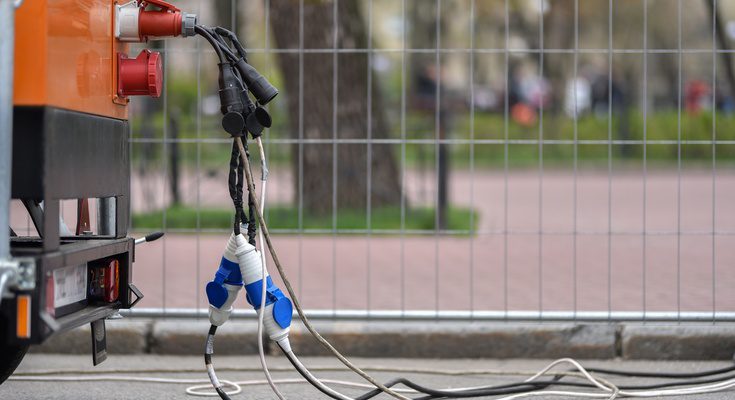 Several cables running out of an orange power generator outside. The generator sits next to a metal fence.