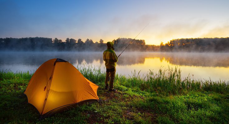 A person is fishing beside a tent at a misty lakeside during sunrise, capturing a peaceful outdoor camping experience.