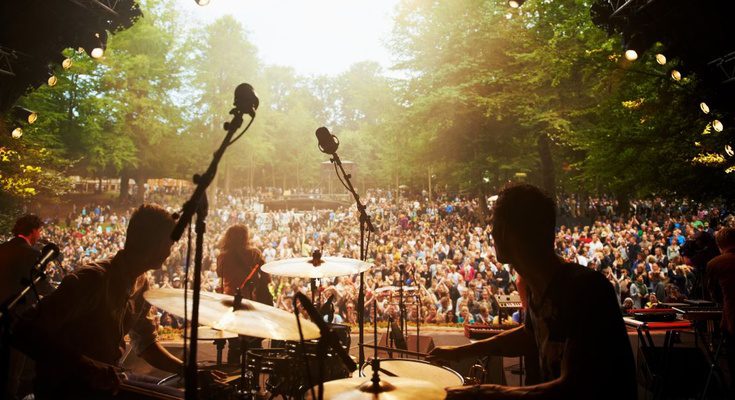 A view looking out from behind a set of drums on a music stage at an outdoor concert with a band and a large audience.