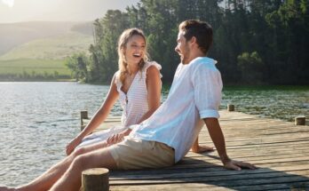 A man and a woman sit next to each other on a dock with rippling water and large trees in the background.