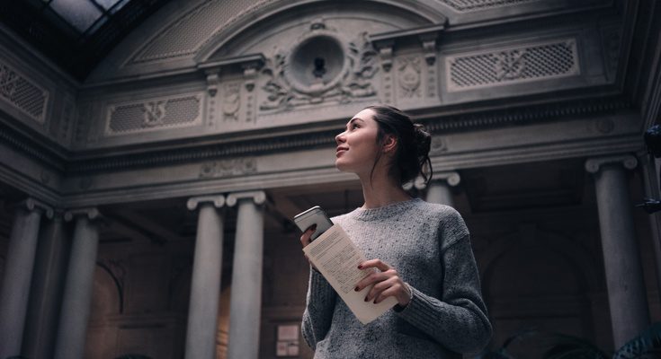 A woman in a gray sweater holds a book and phone while gazing upward inside a grand museum with ornate columns.