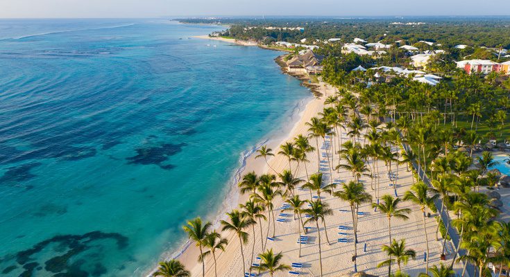 An aerial view shows a beautiful beach with palm trees scattered about and lounge chairs. The ocean is next to the beach.