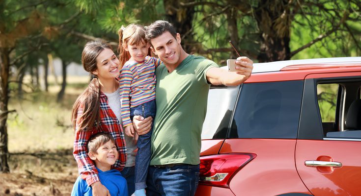 A family of four smiles as they take a selfie together. They stand next to a red car and in front of green trees.