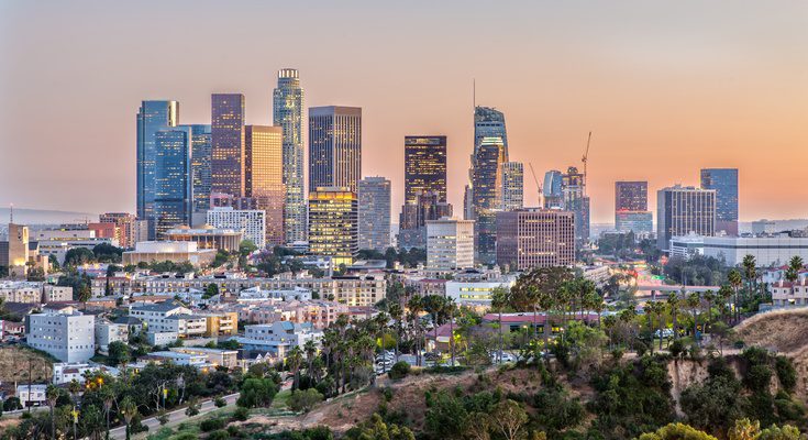 A view of multiple tall buildings with lights on during a sunset, behind multiple smaller buildings and trees.