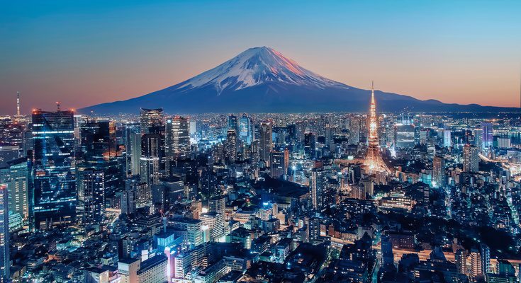 An aerial view of Japan with a large mountain behind the cityscape. The sun is setting and the mountain has a snowcap.